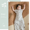 A baby is sleeping in a light gray cotton vest sleeping bag on a woven brown blanket.