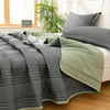 A gray quilted blanket with light green trim is draped over a bed, next to a window.