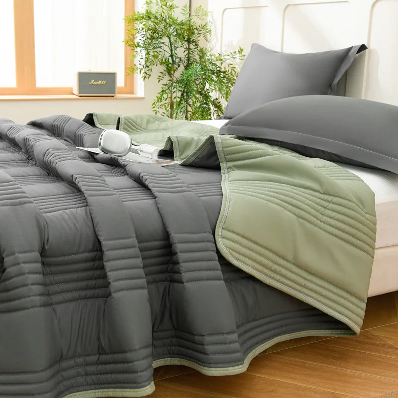 A gray quilted blanket with light green trim is draped over a bed, next to a window.