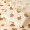 A beige baby swaddle blanket with a light pattern of brown bear footprints is displayed on a textured fabric.