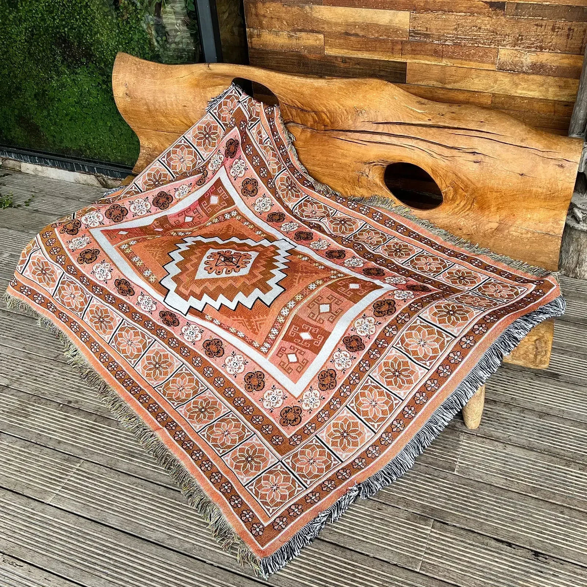 A patterned bohemian geometric throw blanket with earthy tones is displayed on a wooden surface next to a large piece of natu