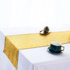 A golden sequin table runner is displayed on a white tablecloth with a small dark pinecone and a teacup.