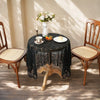 A dark lace tablecloth is set for dining with a white vase of flowers, cups, and pastries on a wooden table.
