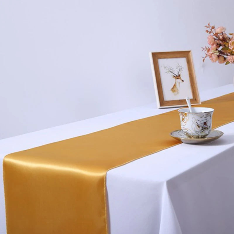 A golden satin table runner draped across a white tablecloth with a small ceramic cup and framed art.