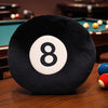 A black soft memory foam ball pillow cushion with the number 8 is resting on a wooden table near a pool table.