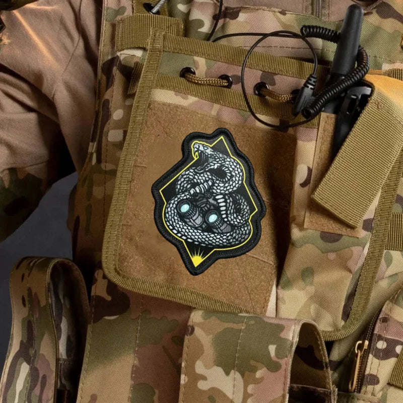 A close-up of a tan camouflage garment featuring a black and yellow snake morale patch sewn onto the fabric.