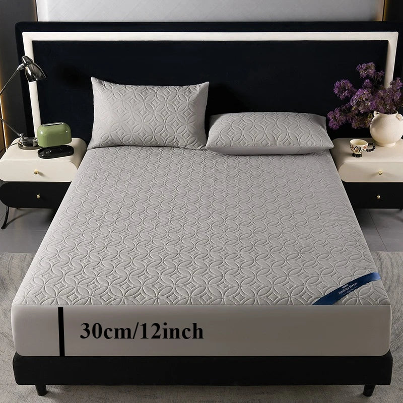 A gray quilted mattress protector is displayed on a bed with dark bedding and pillows.