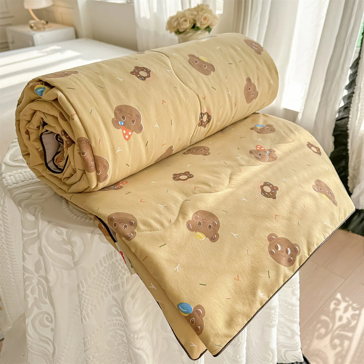 A tan quilt blanket with a pastoral print featuring small brown teddy bears and yellow accents, rolled up on a white bed.