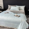 A light blue lightweight cooling blanket is displayed on a bed with gray bedding and pillows.