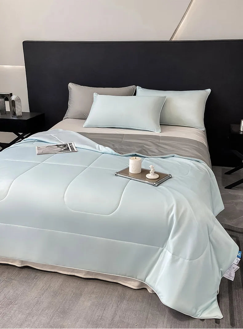 A light blue lightweight cooling blanket is displayed on a bed with gray bedding and pillows.