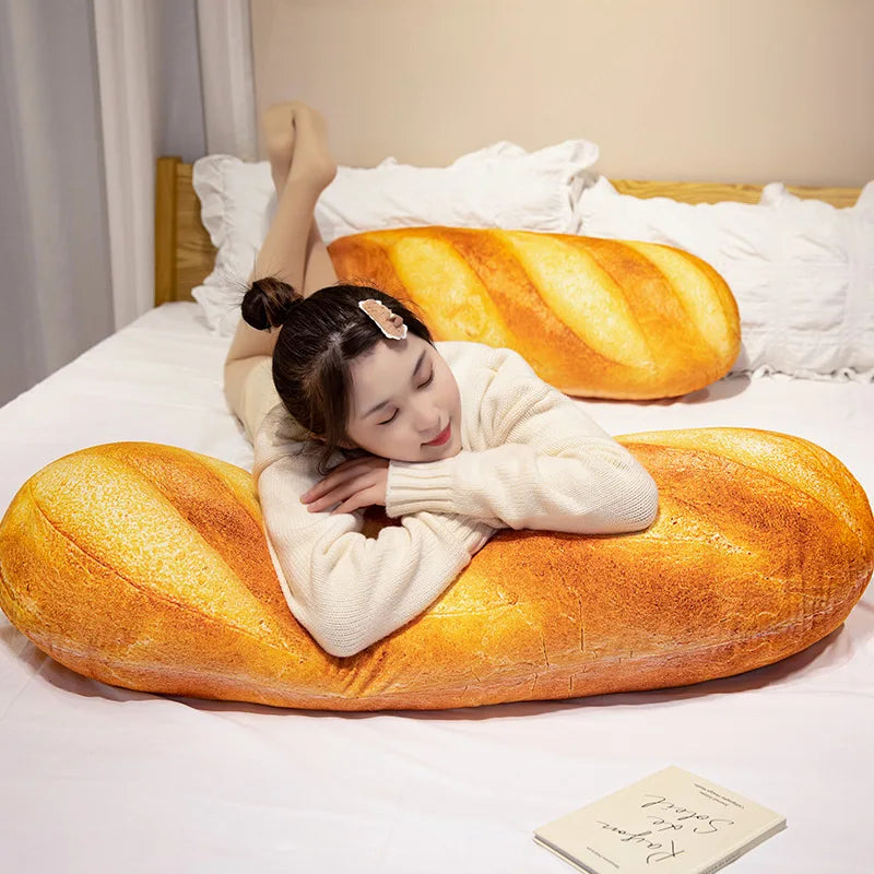 A woman relaxing on a large, golden-brown plush baguette pillow in a bright bedroom setting.