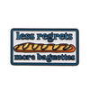 An enamel pin featuring the text less regrets more baguettes in blue and orange lettering.