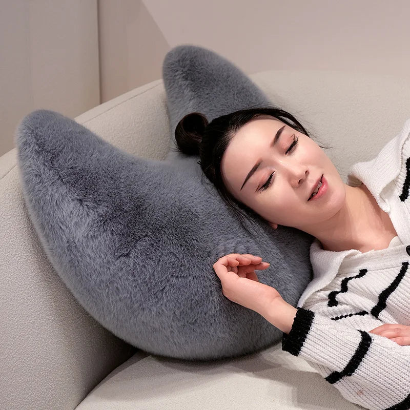 A woman rests her head on a gray moon-shaped throw pillow indoors.