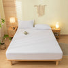 A white fitted sheet set with pillowcases is displayed on a wooden bed in a bright bedroom.
