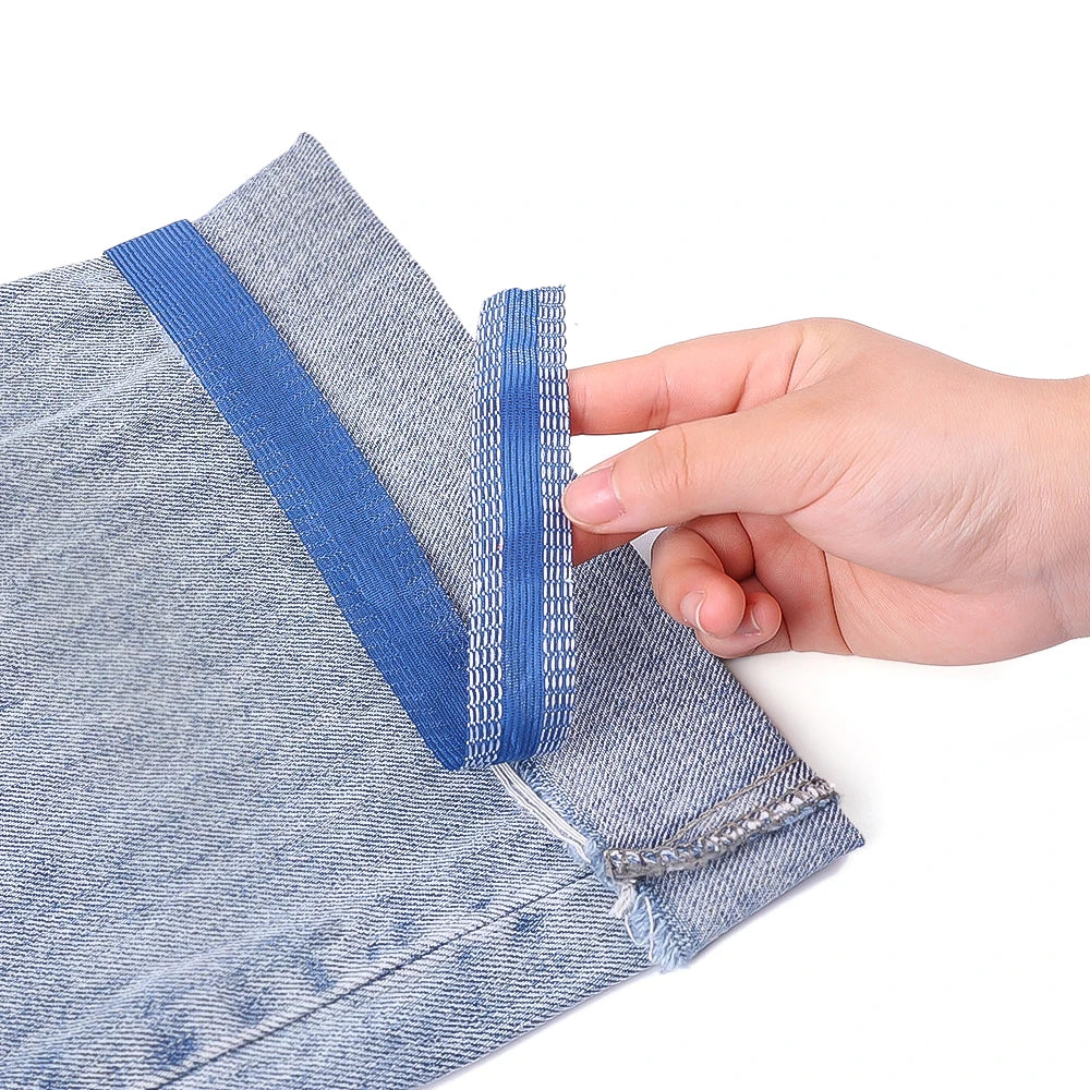 A hand is pulling a blue self-adhesive hem tape from the cuff of light blue denim fabric.