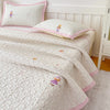 A white floral quilted bedspread with pink trim and small embroidered figures is displayed on a bed.