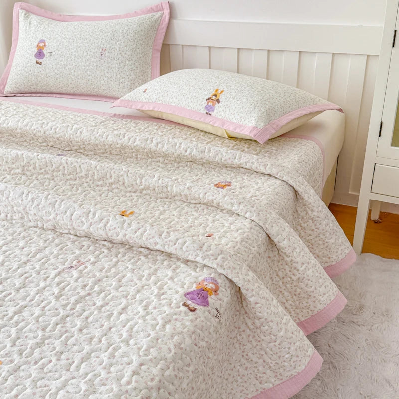 A white floral quilted bedspread with pink trim and small embroidered figures is displayed on a bed.