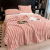 A thick pink fluffy blanket with vertical stripes is displayed on a bed, featuring matching pillows and a throw.