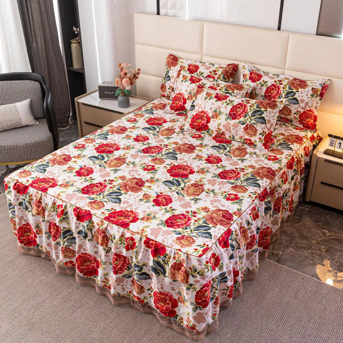 A luxury floral bed skirt in pink and red roses covers a mattress, displayed on a bed with a light upholstered headboard.