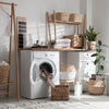 A white washing machine is positioned next to a wooden rack and various woven baskets in a bright interior setting.