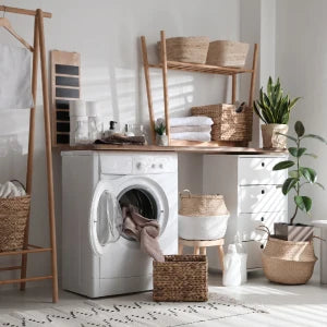 A white washing machine is positioned next to a wooden rack and various woven baskets in a bright interior setting.
