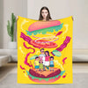 A yellow throw blanket featuring a cartoon illustration of a hamburger and a family standing on the bun.