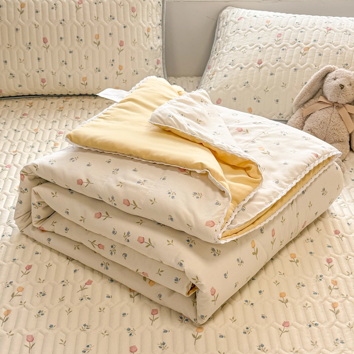 A stack of light beige quilted blankets with a subtle floral pattern resting on a cream-colored quilted surface.