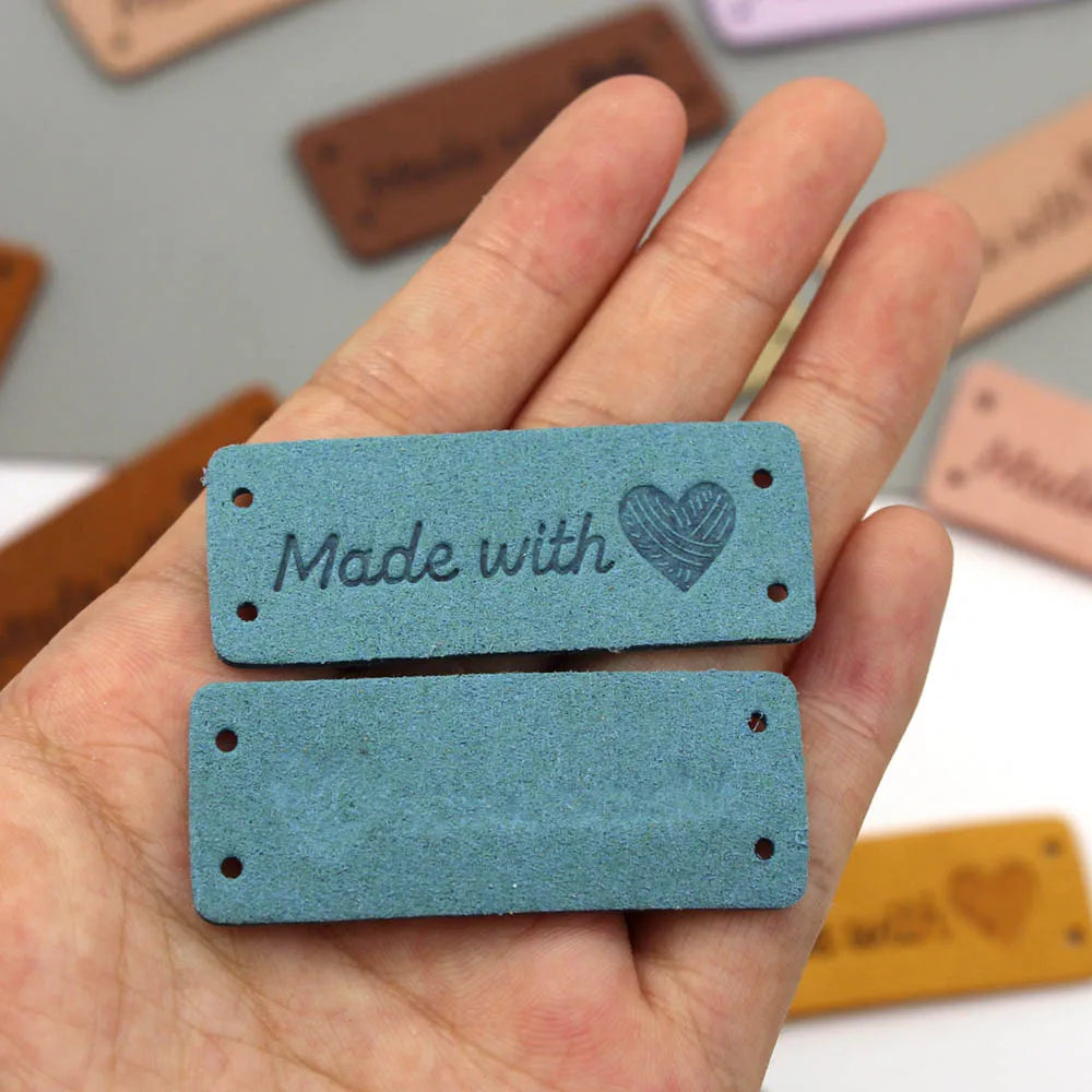 A hand holds two small, rectangular teal leather tags with a heart design on them.