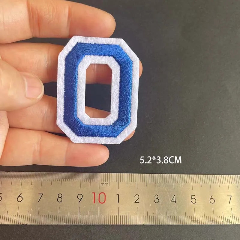 A hand holds a blue and white embroidered iron-on patch shaped like the number zero, measured against a ruler.