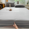 A fitted gray cotton bed sheet is draped on a bed, with a hand touching the fabric.