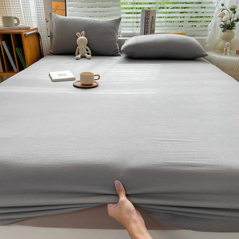 A fitted gray cotton bed sheet is draped on a bed, with a hand touching the fabric.