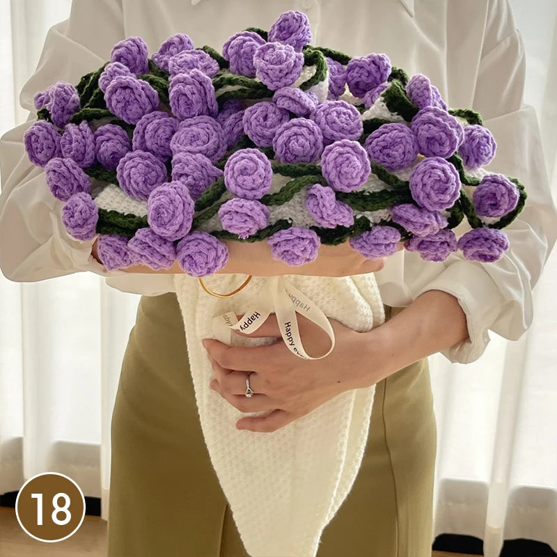 A hand knitted floral rose bouquet arrangement made of purple yarn held by a person wearing a cream blouse and holding a whit