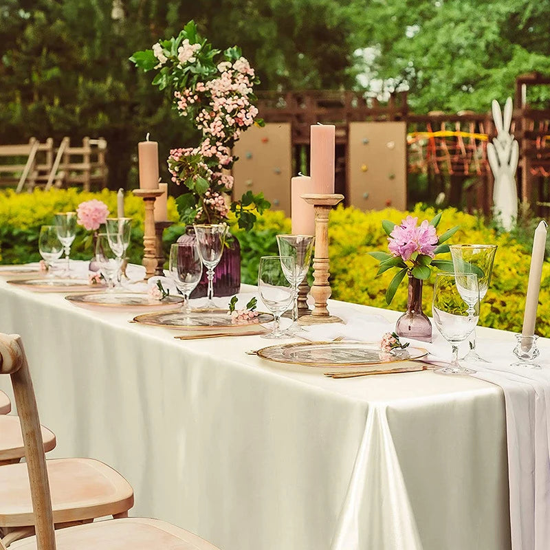 A long table set for an outdoor event featuring a cream satin tablecloth, glassware, candles, and floral arrangements.