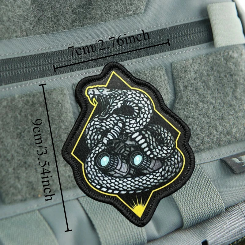 A black and yellow military morale patch featuring a detailed snake design on a gray fabric background.