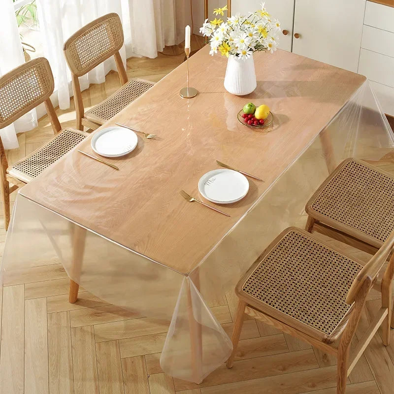 A light wood dining table protected by a clear heat-resistant PVC tablecloth protector with natural wood chairs and floral ce