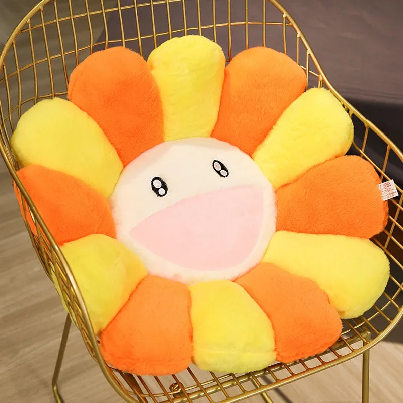 A round, smiling sunflower plush cushion in orange and yellow colors displayed in a gold wire basket.
