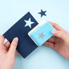 A hand holds a light blue embossing tool against a dark blue card with a star design.