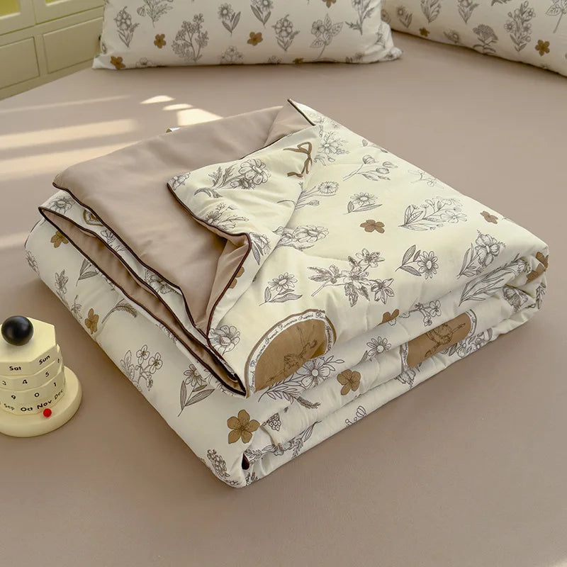 A beige Summer Gauze Cotton quilt folded on a light pink bed with white floral patterns.