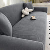 A gray stretch jacquard sofa slipcover with a light-colored decorative pillow and a cup on the seat.