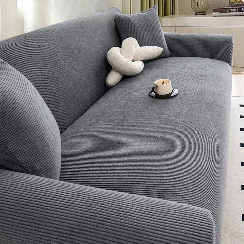 A gray stretch jacquard sofa slipcover with a light-colored decorative pillow and a cup on the seat.