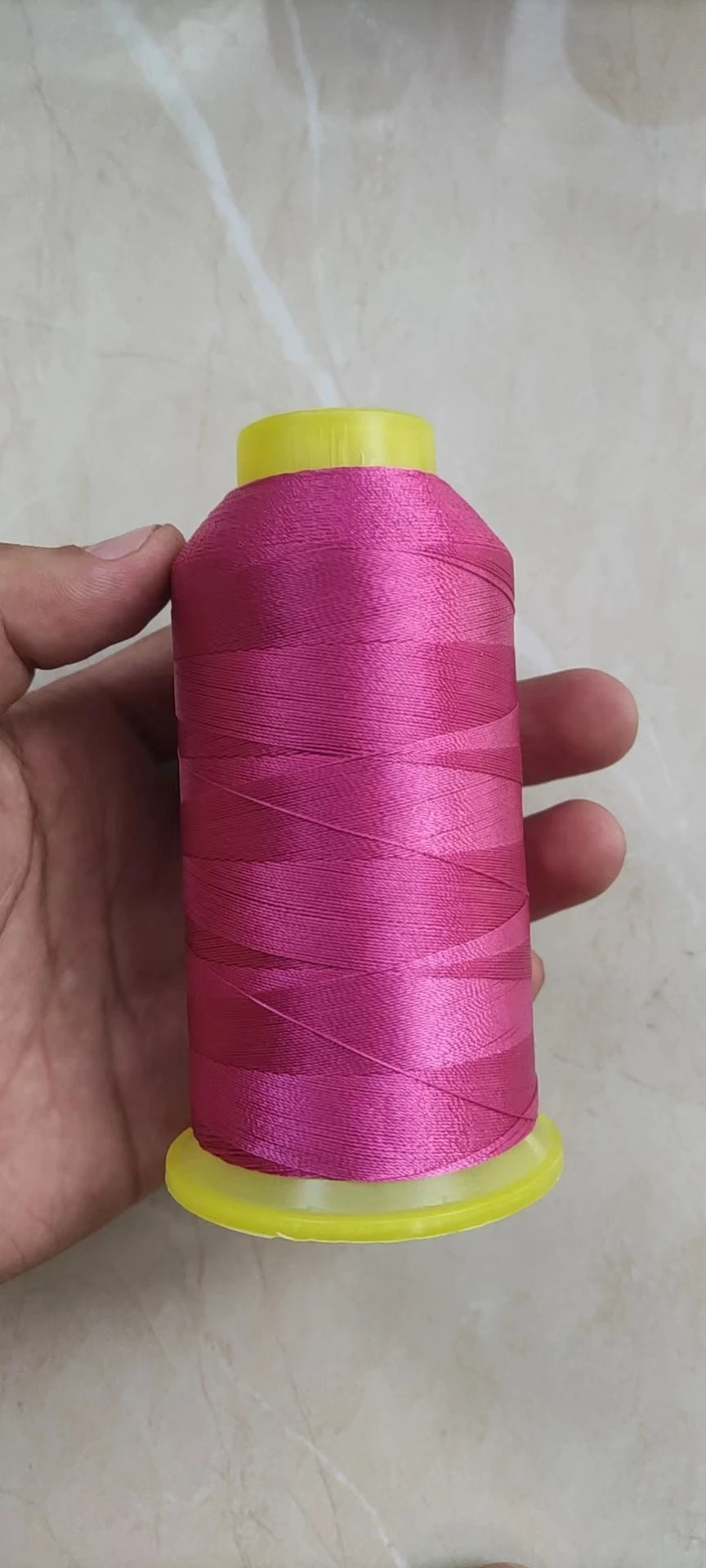 A hand holds a spool of bright pink high tenacity polyester sewing thread with a yellow plastic core.