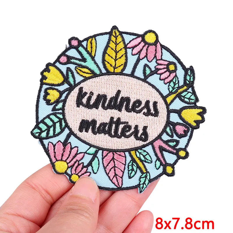 A circular embroidered patch featuring floral elements and the text kindness matters on a light blue background.