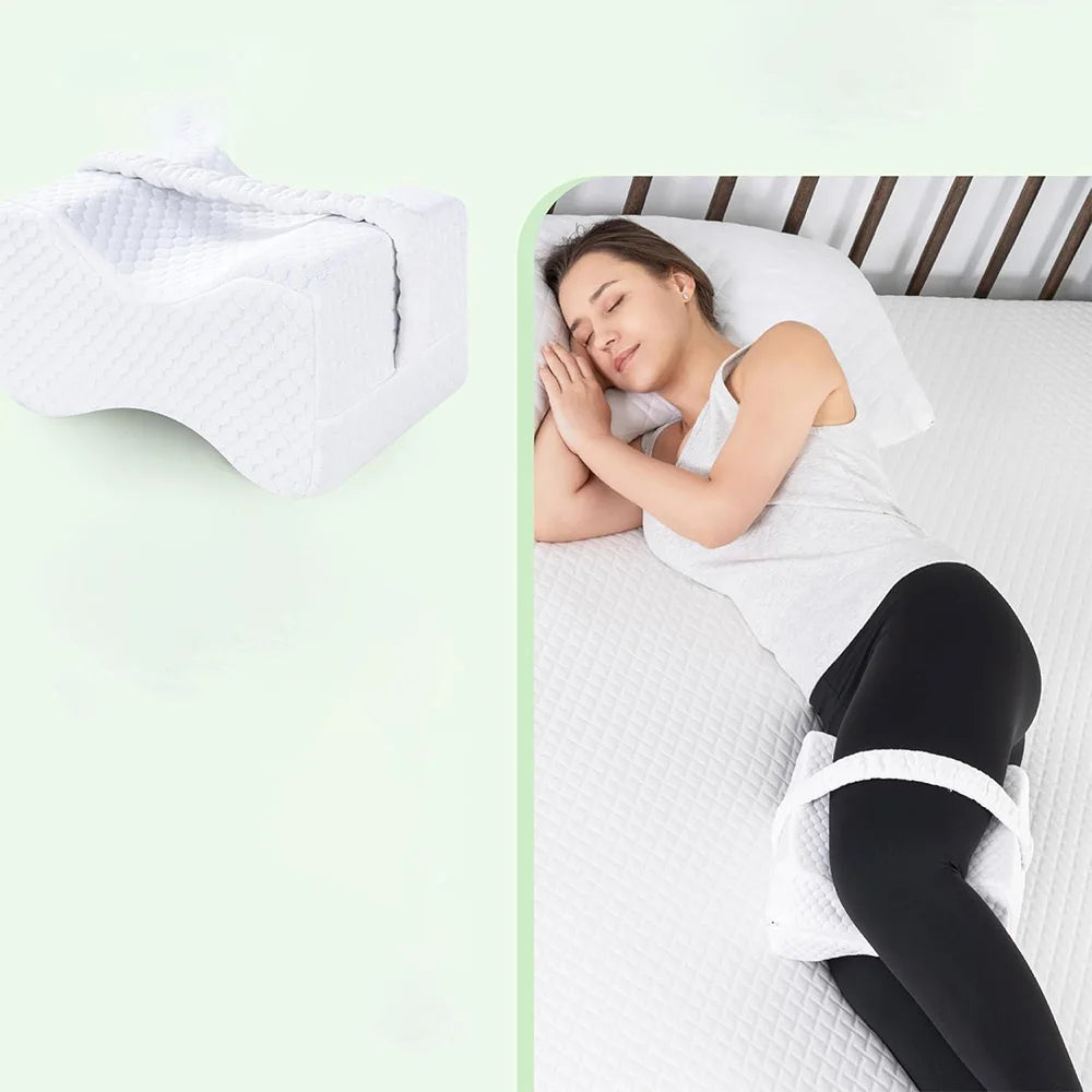 A woman is resting on a bed, using a white memory foam knee pillow with adjustable straps for side sleeping.