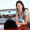 A smiling woman sits in a red convertible, holding a black pillow.