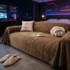 A brown chenille sofa protector with a fishbone pattern is draped over a couch in a dimly lit room.