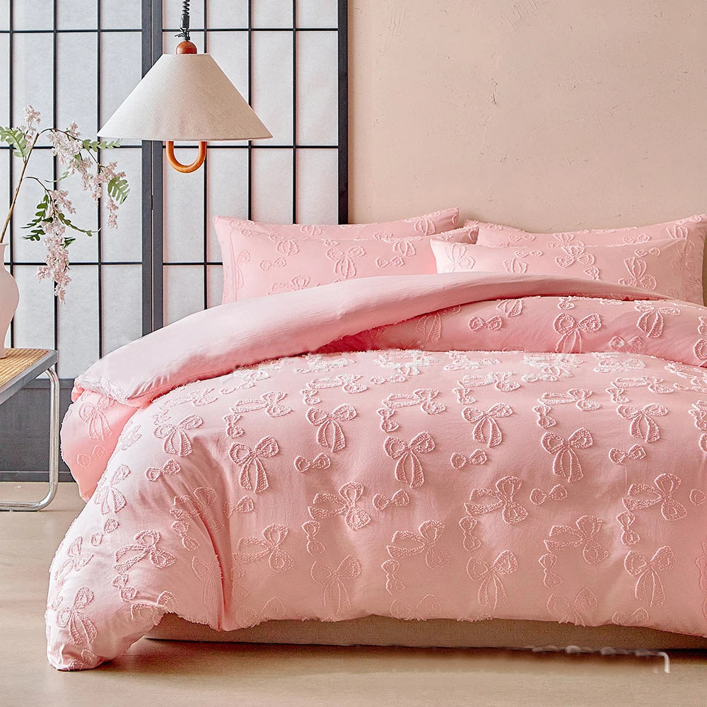 A pink luxury quilt duvet set with white embroidered details is displayed on a bed against a light pink wall.