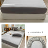 A fitted bed sheet in light gray fabric is shown folded on a bed, with a hand demonstrating its texture.