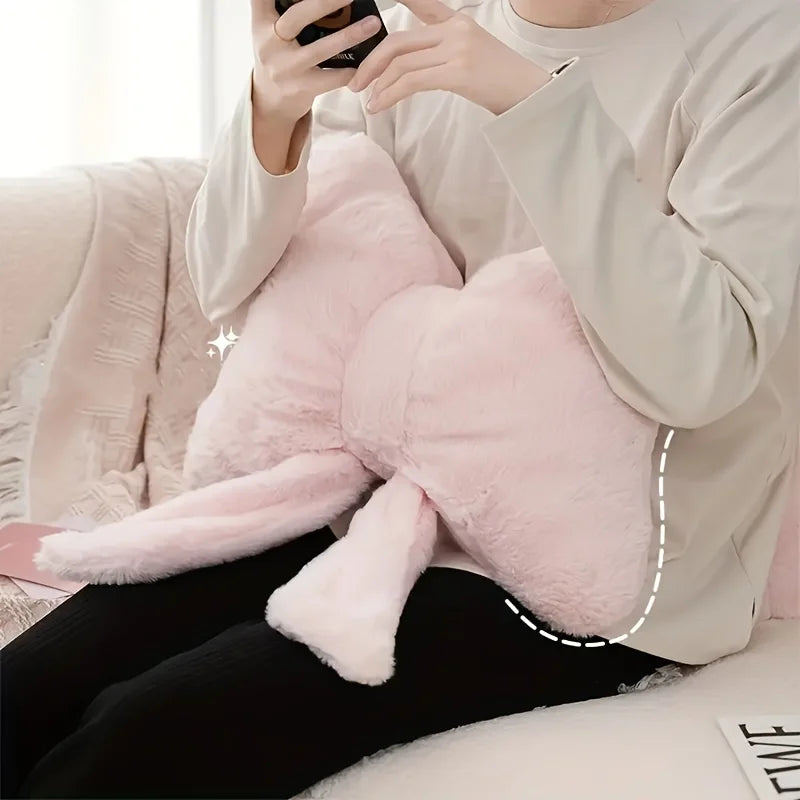 A person is holding a pink fluffy decorative bow pillow in a light beige sweater while sitting down.