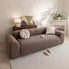 A beige quilted sofa cover with elastic straps is displayed on a light rug in a bright room.