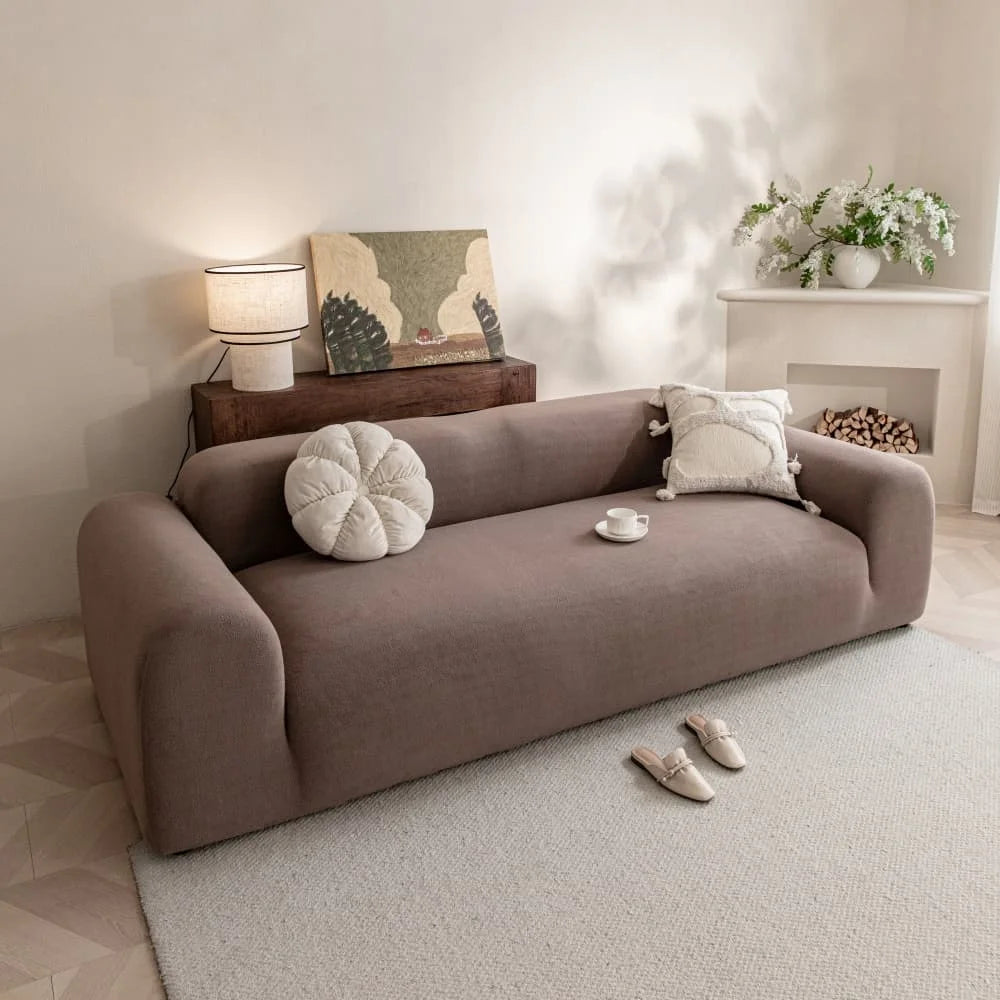 A beige quilted sofa cover with elastic straps is displayed on a light rug in a bright room.